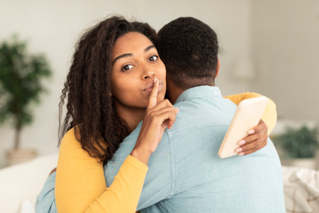 Glad millennial african american wife hugging husband, show silence sign, use smartphone in light living roomの写真素材