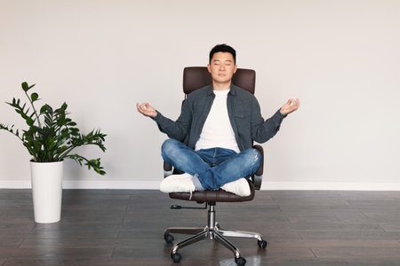 Serious calm adult asian male with closed eyes sits in armchair, meditates, rests from work, enjoys silenceの写真素材
