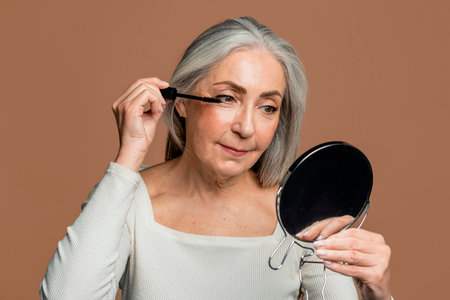 Cheerful senior caucasian female with gray hair applies mascara on eyelashes with brush, looks in mirrorの写真素材