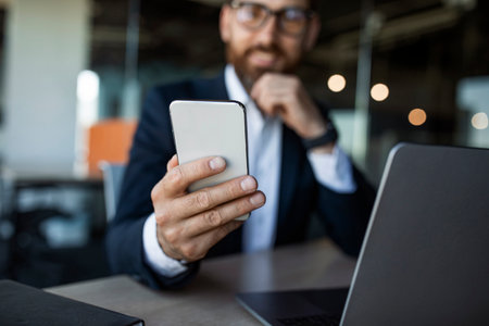 Middle aged businessman sitting at desk and holding cellphone in hand, texting with clients, selective focusの写真素材
