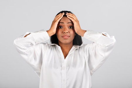 Confused shocked millennial black businesswoman in white blouse holding her hands behind headの写真素材