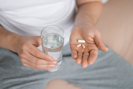 Young caucasian guy suffers from illness, takes pills, holds glass of water, sits on bed in bedroom, top viewの写真素材