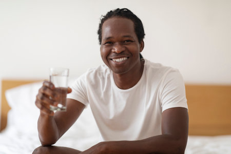 Smiling Black Guy With Glass Of Water Sitting On Bed At Homeの写真素材