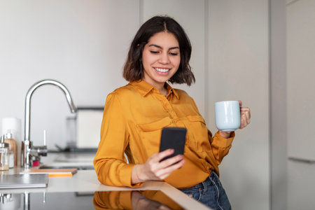 Online Communication. Young Arab Woman Using Smartphone And Drinking Coffee In Kitchenの写真素材