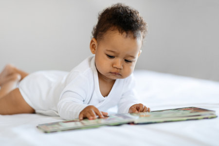 Adorable Black Baby Looking At Pictures In Book While Relaxing In Bedの写真素材