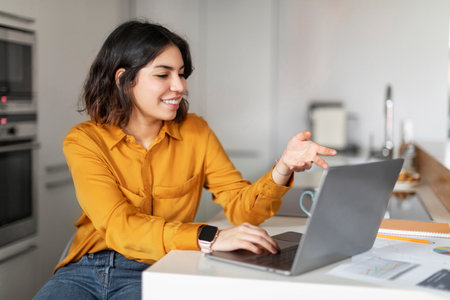 Smiling Young Arab Woman Making Video Call With Laptop In Kitchenの写真素材