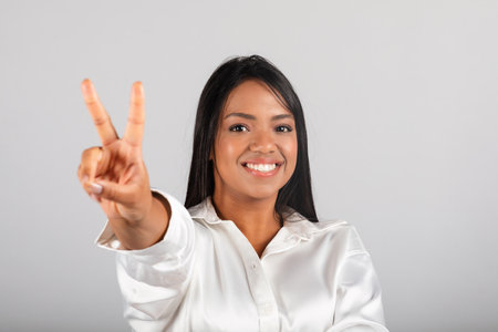 Smiling confident pretty millennial black female in white blouse show peace sign, enjoy good dealの写真素材
