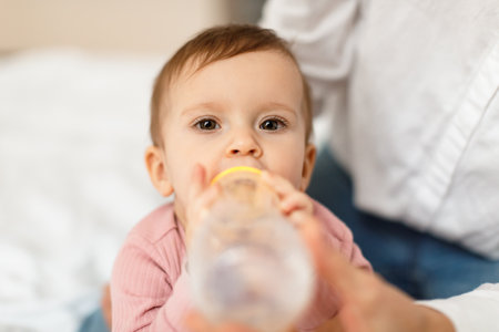 Cute infant baby girl drinking water from bottle, sitting on bed with mommy, thirsty little child enjoying healthy drinkの写真素材