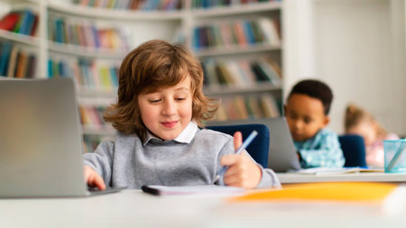 Caucasian boy studying with laptop and writing in notepad, sitting at table in classrom with diverse...