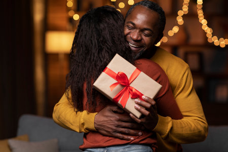 Thankful black man with birthday gift hugging wife at homeの写真素材