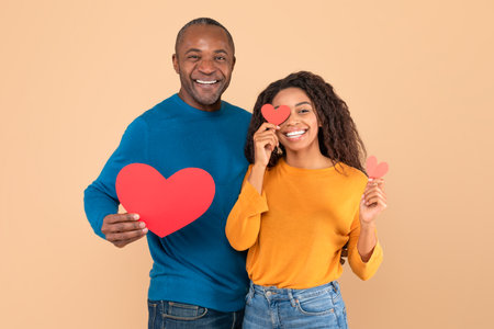 Love is in the air. Romantic black couple with red paper hearts in hands posing over peach studio...