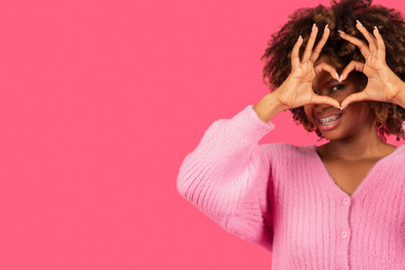 Smiling young african american curly female with braces showing heart sign with hands near eye, have funの写真素材