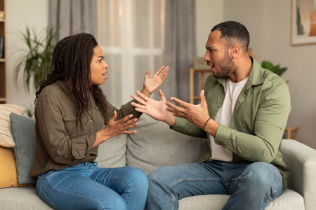 Emotional african american lovers having quarrel, sitting on sofa in front of each other, yelling and gesturingの写真素材