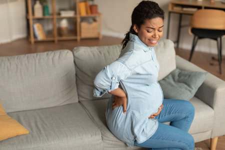 Sad black pregnant lady with big belly suffering from back pain, sitting on sofa in living room, free spaceの写真素材