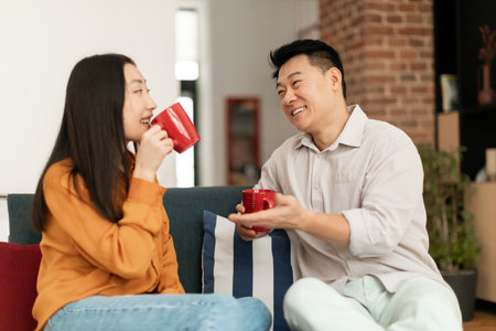 Loving asian spouses drinking coffee and chatting, sitting on sofa at home, enjoying spending time together on weekendの写真素材