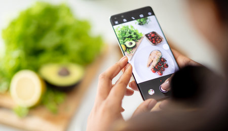 Young african american woman chef makes photo of fresh fish on smartphone screen at table with organic vegetablesの写真素材