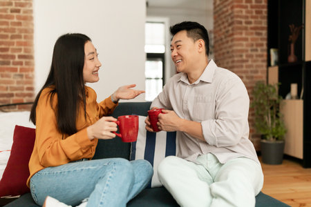 Happy asian spouses sitting on sofa with cups of hot drink and talking, resting in living room interiorの写真素材