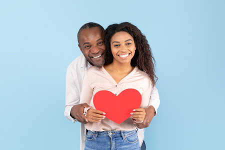 Loving african american couple celebrating their love, black middle aged man and young woman holding big red heartの写真素材
