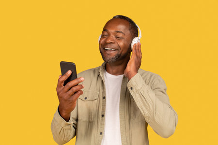 Cheerful relaxed mature african american man in casual and wireless headphones looking at phoneの写真素材