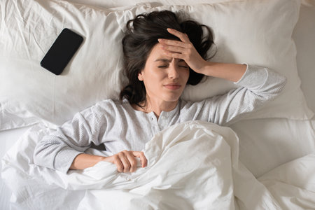 Sad young european woman wakes up, presses her hand to head, suffers from headache, has bad dreamsの写真素材