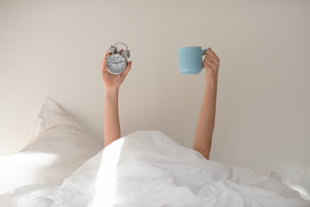 Hands of young european woman under covers with alarm clock, cup of coffee on white bed in bedroom