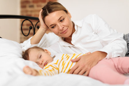 Loving mother hugging sleeping daughter, lying on bed nearby and looking at kid girl