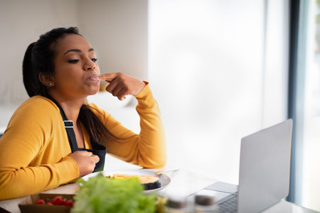 Happy millennial african american woman in apron trying fish dish, licking finger, watching video blog on laptopの写真素材