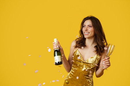 Festive mood concept. Happy woman in dress with champagne bottle and filled glass in hand posing with falling confettiの写真素材