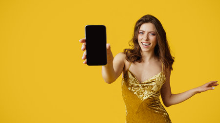 Happy young caucasian woman wearing shiny golden dress and showing cellphone with blank screen, panorama, mockupの写真素材