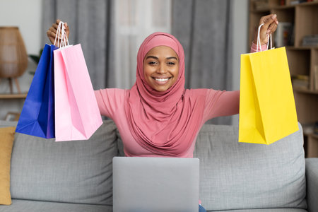 Excited black lady in hijab sitting on sofa with laptop and lifting gift bags above her head, happy to get big discountの写真素材