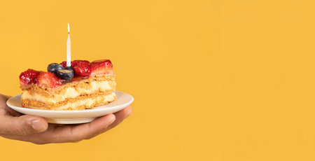 Male hand holding birthday cake with burning candle isolated over yellow backgroundの写真素材