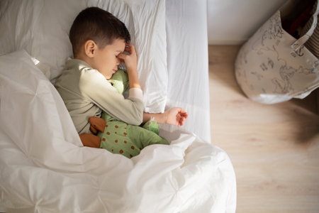 Tired european small kid in pajamas sleeps, wakes up on bed with dinosaur toy in bedroom interiorの写真素材