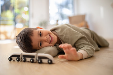 Smiling pensive small european boy in pajamas lies on floor, enjoy relax, free time, plays with cars...