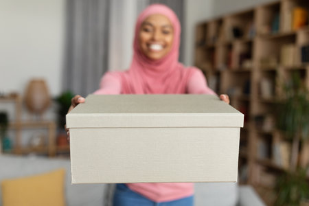 Shopping delivery. Happy black muslim lady stretching cardboard box to camera, standing in living room, selective focusの写真素材