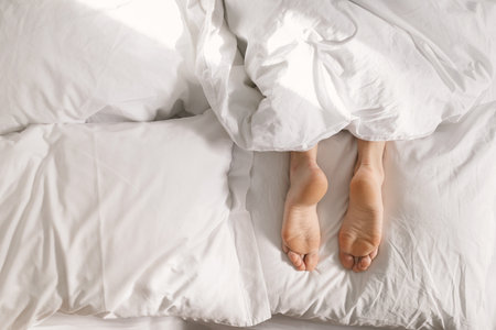 Legs of young european female lies on white comfortable bed under blanket, wakes up, enjoy free time...