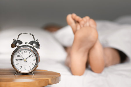 Feet of caucasian millennial lady lies on bed under white blanket, sleep at morning, focus on alarm clock on tableの写真素材