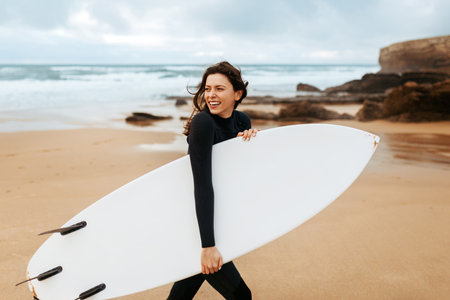 Happy surfer girl walking with board on the sandy beach, looking away and smiling, free spaceの写真素材