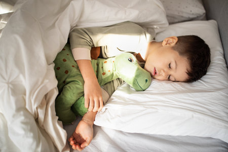 Tired european small kid in pajamas sleeps on bed with dinosaur toy in bedroom, top view, empty spaceの写真素材