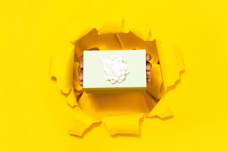 Male hands giving wrapped gift box through torn yellow paper background, closeup, free spaceの写真素材