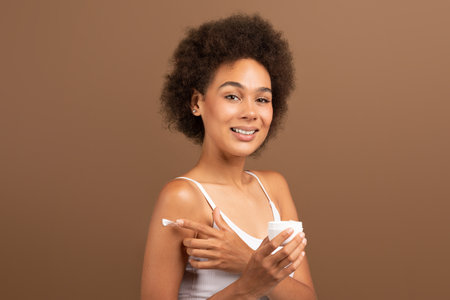 Cheerful young african american curly lady with perfect skin in white top hold jar, applying cream on shoulderの写真素材