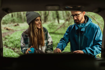 Happy millennial caucasian guy and woman in jackets take stuff out of car trunk in cold forest,...