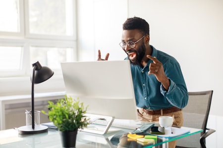 Business success. Emotional black businessman looking at monitor and gesturing yes, exclaiming in joy in officeの写真素材