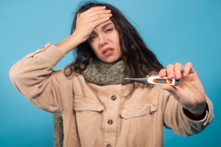 Sad millennial european female in scarf presses hand to forehead, suffers from headache and fever, show thermometerの写真素材