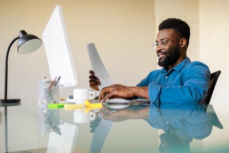 Positive black businessman reading report sitting at workplace in front of computer in officeの写真素材
