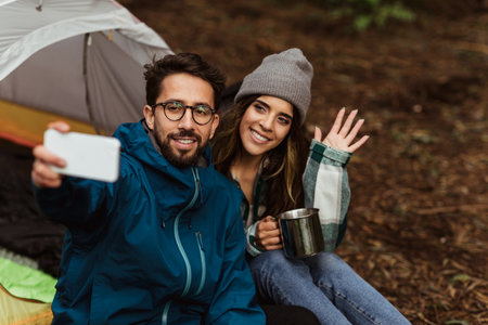 Happy millennial caucasian couple resting in cold forest, enjoy adventure, sit in tent, taking selfie on phoneの写真素材