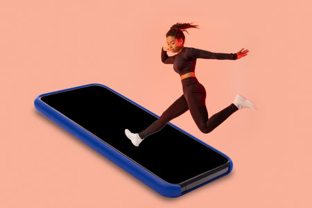 Fitness app. Black woman jumping on smartphone blank screen, having workout on peach studio background, mockupの写真素材