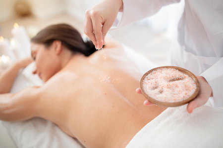 Masseuse applying mineral salt body scrub on back of young womanの写真素材