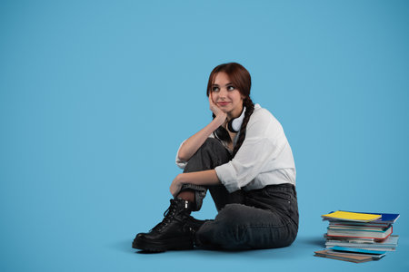 Smiling pensive european teenager girl student with pigtails sitting on floor with books, think, isolated on blue studio background. Break from study, education and homework, choice, ad and offerの写真素材