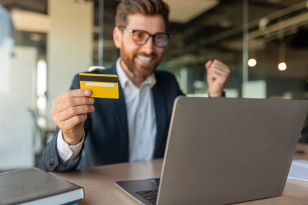 Overjoyed businessman making success gesture, holding credit card in front of laptop sitting at...