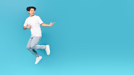 Korean Teen Guy Jumping Pointing Fingers Aside Over Blue Backgroundの写真素材